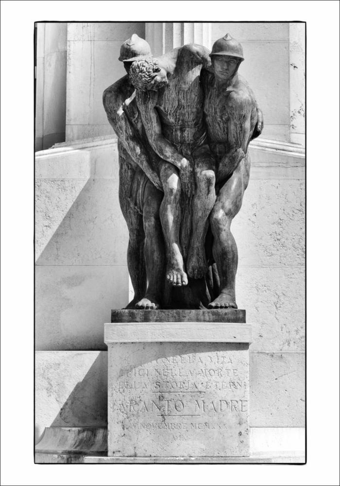 L'immagine digitale in bianco e nero cattura una parte dell'imponente Monumento ai Caduti di Taranto. Inaugurato nel novembre del 1930 alla presenza del re Vittorio Emanuele III, è il più grande monumento del Borgo Nuovo collocato al centro di Piazza della Vittoria, a pochi passi dal corso principale cittadino. Ideato e disegnato dallo scultore originario di Taranto Francesco Paolo Como, è di tipo "a cippo", realizzato in pietra calcarea di Trani con tre gruppi di statue in bronzo. Nella parte superiore, un gruppo di soldati è accompagnato da Atena, dea della guerra, e Vittoria, la dea della vittoria in battaglia; in quella inferiore, un gruppo, denominato Aquilifero, rappresenta un soldato nell'atto di scagliare in volo un'aquila, sull'altro lato, l'ultimo gruppo raffi...