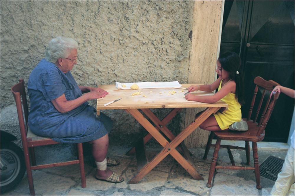 La fotografia è parte della ricca raccolta fotografica “Puglia per tutte le Stagioni” vol. 2, prodotta dall'Assessorato al Turismo e Industria Alberghiera della Regione Puglia e creata per raccontare la Puglia attraverso immagini diverse suddivise in 5 settori principali: Città e Paesi, Settimana Santa, Territorio, Feste – Sagre – Tradizioni, Mercati – Prodotti – Cucina. Lo scatto mostra un momento che facilmente si può osservare per i vicoli dei centri storici pugliesi, la preparazione delle orecchiette fatte a mano. L'orecchietta è in assoluto la “regina” della pasta fresca della tradizione culinaria pugliese anche se è conosciuta con nomi differenti a seconda della località di produzione. In Terra di Bari è nota come “strasc'nat” in quanto la pasta viene trascinata s...
