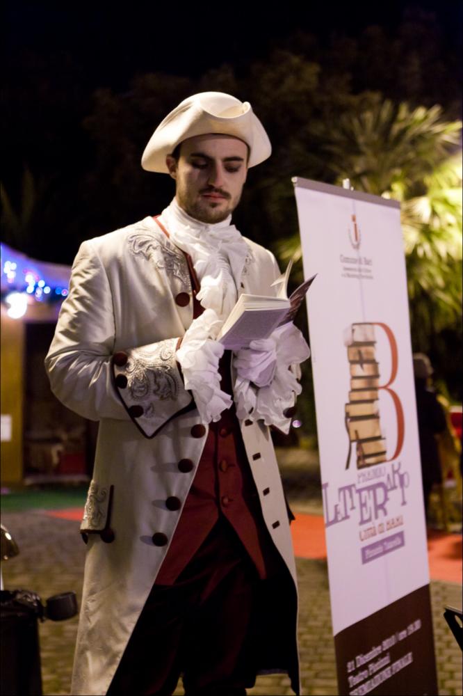 L'immagine digitale documenta una delle azioni di teatro urbano, messe in scena dall'Associazione Fibre Parallele, durante l'evento "Premio Letterario Città di Bari", dell'edizione 2010. La fotografia riprende l'azione teatrale nel più grande parco cittadino, Parco Due Giugno, con l'attore in procinto di leggere le pagine del libro “Otto personaggi in cerca (con autore)” di Björn Larsson, vincitore del Premio Speciale per la Narrativa. Il mondo delle scienze rimane per lo scrittore l'unico mondo possibile ancora da scoprire. E' qui che parte il viaggio dei suoi personaggi, che si mettono in gioco pur di inseguire la loro ricerca. L'edizione 2010 del Premio, alla sua XIII edizione, ebbe il pregio di esaltare le opere letterarie degli scrittori in finale, coinvolgendo att...