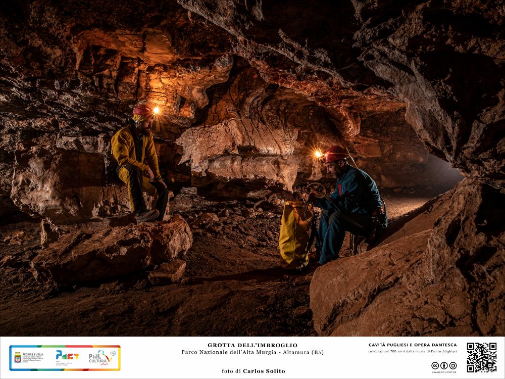 I nostri speleologi riprendendo fiato all’interno della Grotta dell’Imbroglio, una delle grotte di maggiore estensione ubicate nel settore orientale del Pulo di Altamura, che si estende per  diverse decine di metri. Il soffitto è decorato da accentuati fenomeni di corrosione ed evidenze di concrezionamento fossile e il pavimento è caratterizzato da terreno di colore rosso, massi calcarei crollati sia dal soffitto e che dalle pareti. 
L’immagine digitale fa parte della raccolta di fotografie prodotte da Carlos Solito per il Progetto digitale “Sillabario sotterraneo – Viaggio dantesco nelle Grotte di Puglia” – realizzato nell’ambito della LEGGE REGIONALE 30 dicembre 2020 - art. 39 “Cavità pugliesi e opera dantesca - Celebrazioni 700 anni dalla morte di Dante Alighieri”.