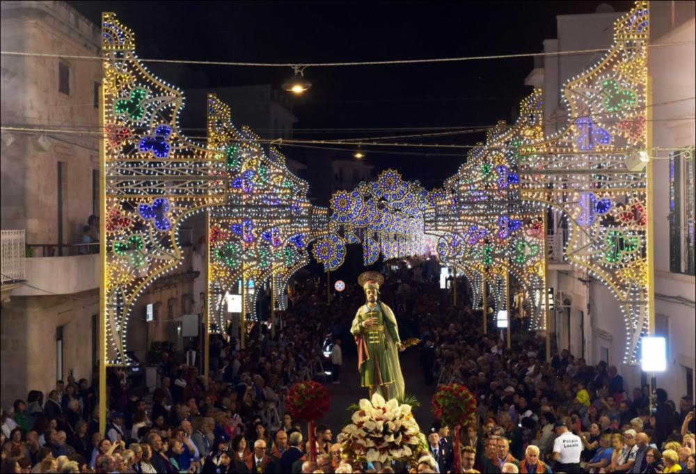 Lo scatto cattura il solenne passaggio dell’imponente statua di San Damiano, che sfila tra le vie cittadine illuminate da sfavillanti luminarie e ornate di corone floreali, mentre una folla di fedeli in festa accompagna il corteo con devozione e gioia.
La Festa Patronale dei Santi Medici Cosma e Damiano, istituita il 12 aprile 1854, si celebra ad Alberobello, in provincia di Bari, dal 25 al 28 settembre.
Il 25 settembre si apre con la tradizionale fiera dell’antico bestiame e degli utensili agricoli. Il 26 settembre si celebra la festa liturgica. Il 27 settembre è dedicato ai pellegrini: la giornata inizia con la prima Messa alle ore 4.00, seguita dalla solenne processione mattutina alle ore 11.00. Il 28 settembre si concludono i festeggiamenti con ulteriori celebrazion...