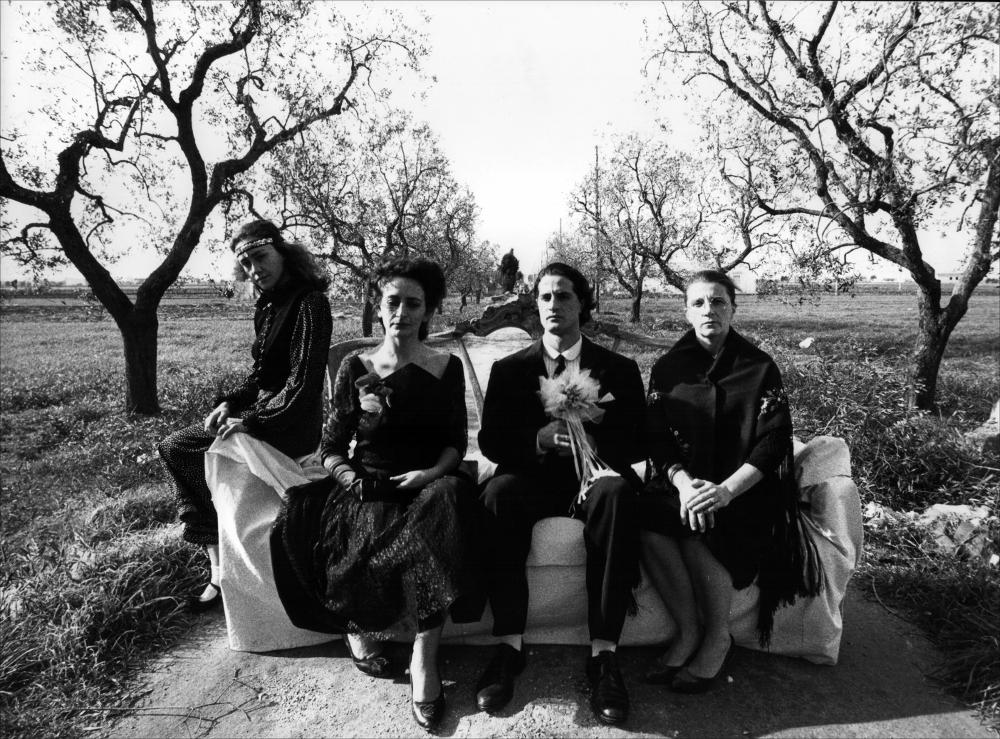 La fotografia in bianco e nero cattura un momento della rappresentazione teatrale “Amori”, messa in scena nel 1988 dal Teatro Koreja, nell'ambito della rassegna annuale “Aradeo e i Teatri”, un appuntamento con il teatro sperimentale che la compagnia nata nel 1983 nel piccolo borgo salentino di Aradeo porta avanti sin dall'anno della sua fondazione. “Amori” trae ispirazione dal dramma “Le tre sorelle” di Anton Checov del 1900, immerso nei vizi e nelle dinamiche spesso soffocanti della realtà meridionale. Accomunate dallo stesso destino di vivere insieme, nella stessa casa, in un paese dove non succede niente, le tre sorelle Caterina, Maria e Teresa non possono far altro che raccontare al loro amico "del cuore" i loro segreti, le passioni e la loro voglia di evadere da qu...