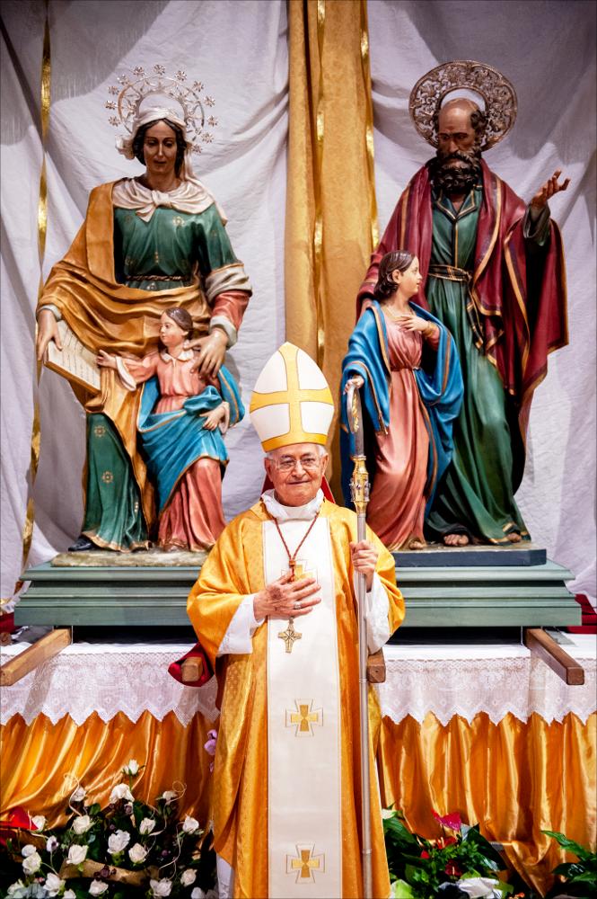 La fotografia ritrae il Vescovo mentre sosta davanti alle statue votive dei patroni di Vernole, poste in posizione d’onore all’interno della chiesa o durante un momento celebrativo. 
Sant’Anna è raffigurata con un libro aperto tra le mani, simbolo dell’educazione alla fede e della trasmissione delle Sacre Scritture alla figlia Maria. Talvolta tiene accanto a sé la giovane Vergine, a indicare il suo ruolo di guida e maestra.
San Gioacchino è rappresentato con atteggiamento paterno e con attributi che richiamano la sua giustizia e rettitudine; accanto a sé la giovane Vergine, a indicare il suo ruolo di guida.<div><br>La festa patronale di Vernole, dedicata a Santi Gioacchino e Anna, si celebra 25-26 luglio, con radici che risalgono al 1681, anno in cui fu completata la ca...