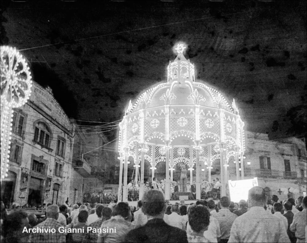 Fotografia storica del 1970. Scatto recuperato e digitalizzato dall’Archivio Gianni Pansini, ritrae la banda del paese all’interno della cassa armonica, impegnata nell’esecuzione di brani musicali durante la festa patronale. L’immagine cattura l’energia e la partecipazione del pubblico, con la piazza animata dai suoni della musica e dall’atmosfera festiva tipica delle celebrazioni patronali canosine.
<div><br>La festa patronale di San Sabino si celebra ogni anno il 1° agosto a Canosa di Puglia e rappresenta un momento centrale della vita culturale e religiosa della città. La ricorrenza rievoca la traslazione del corpo del santo dalla Basilica di San Pietro alla basilica a lui dedicata, un evento che unisce fede, storia e tradizione popolare.
La festa è caratterizzata da...
