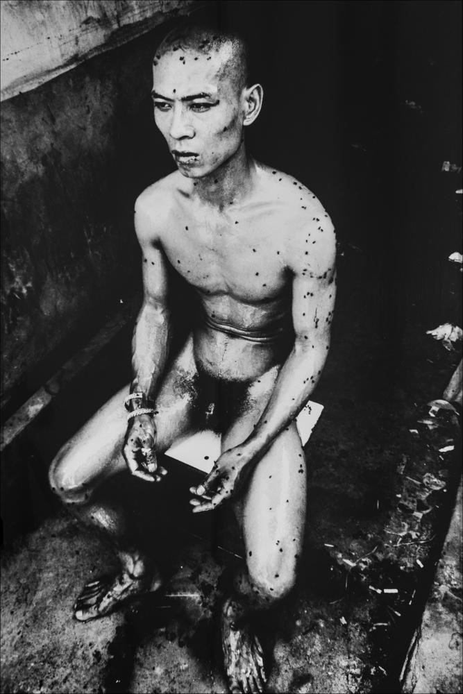 L’immagine presenta l’opera fotografica tratta dalla performance “12 m2” realizzata dall’artista cinese Zhang Huan nel 1994. Agli inizi della sua carriera, Zhang Huan viveva in una comunità di artisti in un quartiere molto povero e degradato nella periferia di Pechino, e spesso metteva in scena performance durante le quali testava i limiti del disagio della propria resistenza fisica.
L’immagine di “12 m2” è quella di un uomo seduto, con la fronte alta, il dorso eretto e le mani raccolte tra l’anca e il ginocchio. Ciò che suscita sconcerto è che il corpo, completamente nudo e ricoperto da una sostanza viscida, è un ricettacolo di sciami di mosche e insetti che si fissano dappertutto, sulla bocca e sulle orecchie, rimanendo invischiati su tutta la pelle. Immerso nello spa...