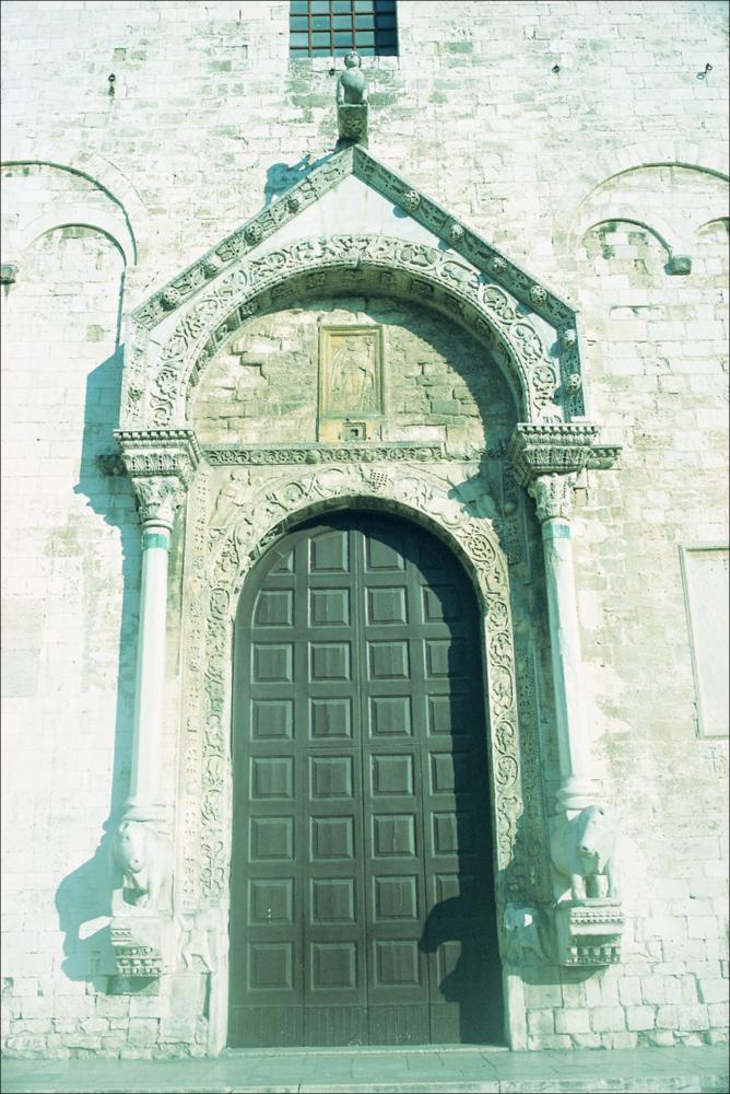 La fotografia è parte della ricca raccolta fotografica “Puglia per tutte le Stagioni” vol. 2, prodotta dall'Assessorato al Turismo e Industria Alberghiera della Regione Puglia, per raccontare la Puglia attraverso immagini suddivise in 5 settori principali: Città e Paesi, Settimana Santa, Territorio, Feste – Sagre – Tradizioni, Mercati – Prodotti – Cucina. La fotografia mostra un primo piano del portale della Basilica di san Nicola di Bari, il più importante edificio di culto della città e un esempio del romanico pugliese, voluto dall'abate Elia per ospitare le reliquie del Santo trafugate a Myra, Turchia, durante una spedizione di 62 marinai. Fondata nel 1087 e conclusa nel 1089, la Basilica fu costruita sul precedente Catapano bizantino, che secondo molti giustifichere...
