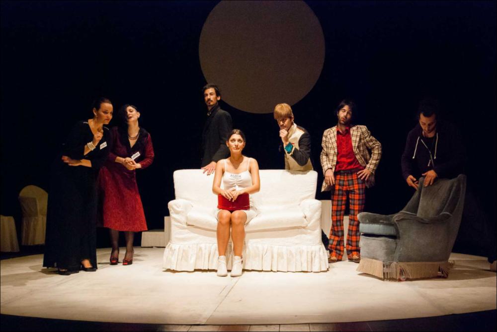 L'immagine in oggetto, fotografia digitale ad opera di Angela Chezzi, cattura un momento dello spettacolo “Il Matrimonio”, per la regia di Salvatore Tramacere, tratto dall'opera teatrale del 1842 di Nikolaj Vasil'evič Gogol', satira incentrata su una giovane donna, Agafija, corteggiata da quattro scapoli, ognuno con le sue eccentricità. Gli stessi personaggi vengono catapultati da Tramacere nel nostro tempo, portando con sé i lati più buffi e divertenti. Il palcoscenico ricorda un set di una trasmissione televisiva, con un presentatore che guida uomini e donne nella ricerca del partner, ma tutto risulta finto e lo schermo allontana ancor più i protagonisti, lasciandoli soli con le proprie inquietudini, aumentando così la distanza tra l'essere e l'apparire. Per far sposa...