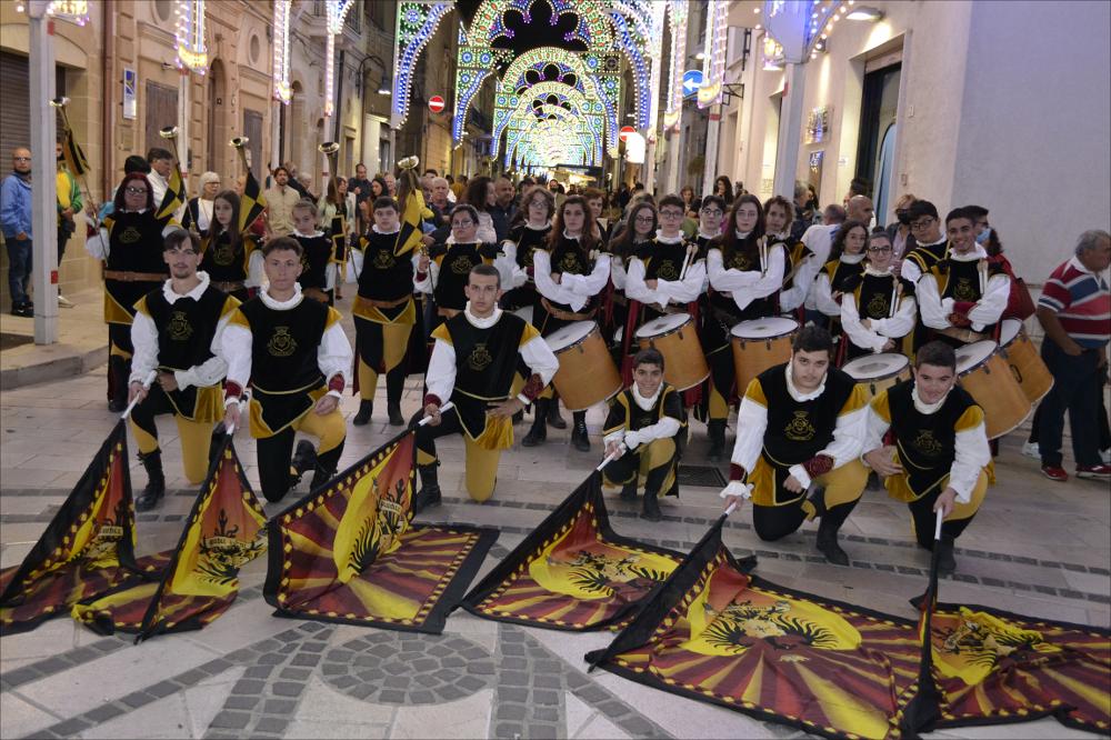 <div class="ql-editor"><div><span>Scatto che ritrae gli </span><strong>sbandieratori in posa</strong><span> nella loro composizione completa durante la Festa Patronale di Villa Castelli nel 2021. I figuranti, vestiti con i tradizionali costumi storici, animano le strade del paese con movimenti sincronizzati e lanci di bandiere dai colori vivaci, creando un effetto scenico e spettacolare. </span>La performance degli sbandieratori unisce <strong>abilità, precisione e ritmo</strong>, trasformando le piazze in palcoscenici vivi e mantenendo un legame diretto con le tradizioni storiche e popolari della comunità.</div><div><br></div><div>La Festa Patronale di <strong>Villa Castelli</strong> si svolge ogni anno dall’1 al 3 ottobre ed è uno degli appuntamen...