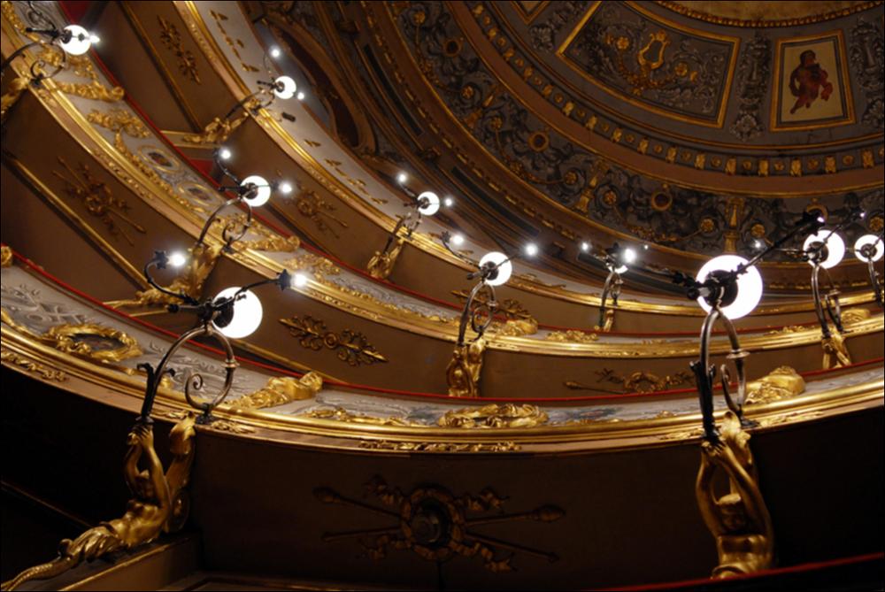 L'immagine è parte della campagna fotografica realizzata dalla fotografa Margherita Mirabella per Teatro Pubblico Pugliese, che con i suoi scatti ha ripreso le eleganti architetture e i rifiniti dettagli di alcuni dei più bei teatri pugliesi e di alcuni spettacoli teatrali portati in scena in questi ultimi anni. Lo scatto immortala un particolare della zona dei palchi, all'interno di Teatro Curci di Barletta, importante contenitore culturale cittadino intitolato al compositore Giuseppe Curci situato in pieno centro. L'aspetto attuale è quello del nuovo teatro, costruito interamente sul precedente e più antico teatro di San Ferdinando, per volontà del comune, che lo acquistò ordinandone l'abbattimento dopo il suo crollo parziale. I lavori iniziarono nel 1868 con la direz...