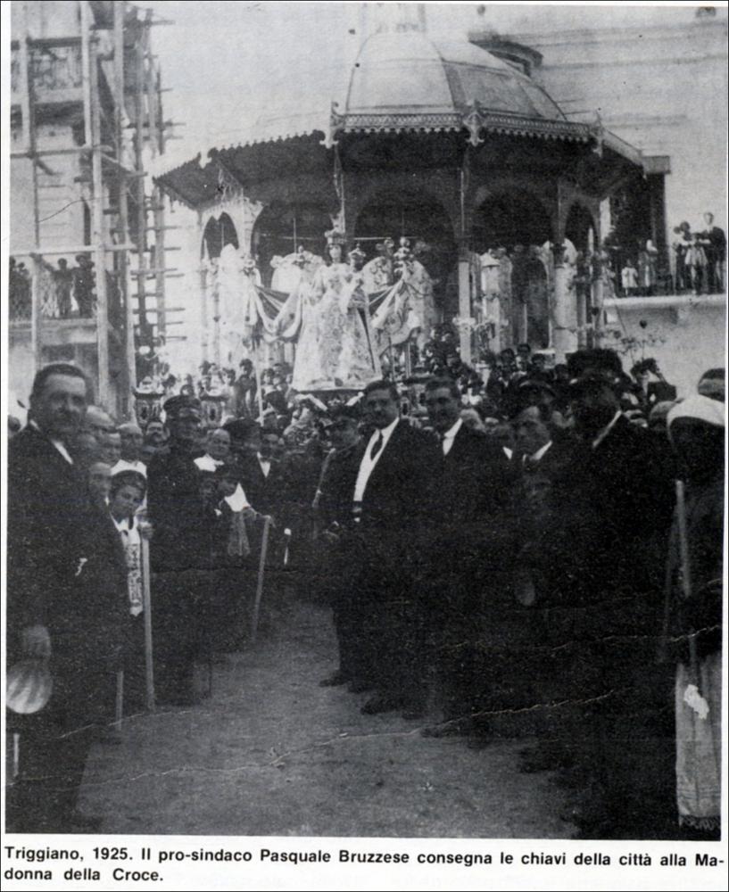 La fotografia storica del 1925 immortala un momento altamente simbolico: il prosindaco Pasquale Bruzzese consegna solennemente le chiavi della città di Triggiano alla Madonna della Croce. Attorno alla maestosa statua della Vergine, allestita per la cerimonia e per i festeggiamenti in Suo onore, si raccoglie una folla numerosa di fedeli, testimoni di un gesto carico di devozione e significato civico.
Le origini della devozione alla Madonna della Croce risalgono al XVI secolo, quando, fuori dall’antico abitato, venne edificata un’edicola sacra decorata con un’immagine “miracolosa” ad opera dei cosiddetti “homini de fora”, ossia contadini e braccianti che ogni giorno passavano di lì per andare a lavorare nei campi.
Una guarigione ritenuta miracolosa, avvenuta proprio nei p...