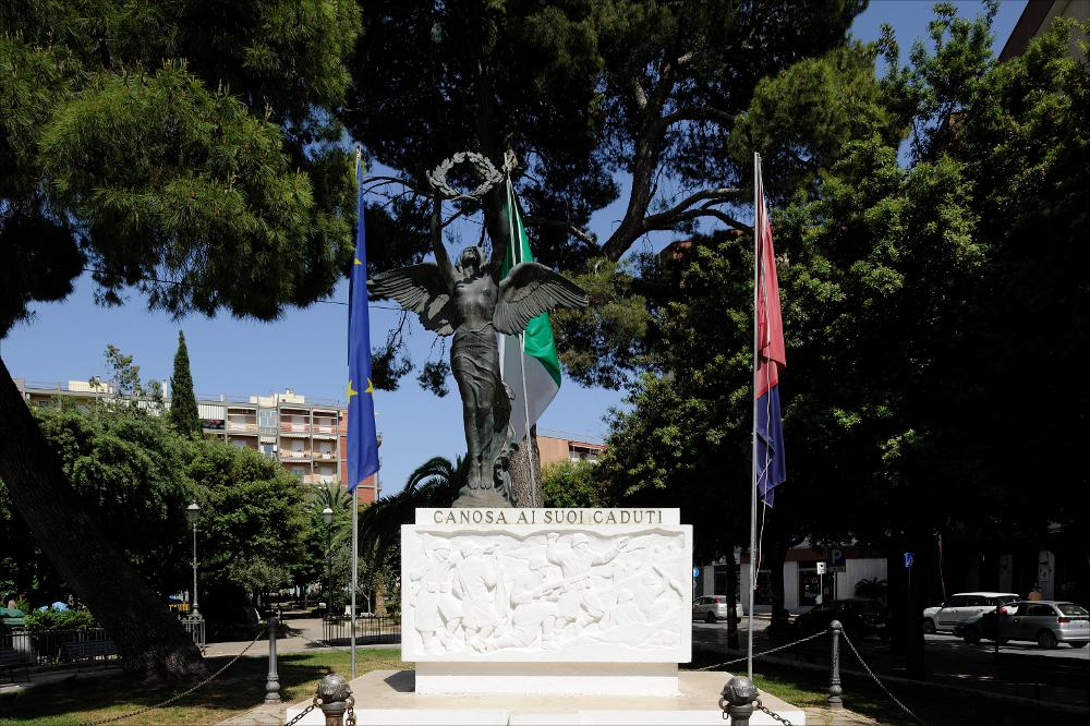 L'immagine digitale mostra il Monumento ai Caduti a Canosa di Puglia. Su un basamento istoriato con immagini di soldati immortalati durante un combattimento vi è l'allegoria della Vittoria come donna alata che innalza una corona d'alloro. È possibile risalire all'iter che ha portato alla costruzione del monumento partendo dalla lettura del carteggio custodito presso l'Archivio Storico Comunale di Canosa di Puglia. Rispondendo ad un bando pubblico emanato dal Comune di Canosa sulla "Gazzetta di Puglia", Francesco Pinto si propone come scultore per la realizzazione del monumento. In una lettera, datata 4 settembre 1924, Pinto dichiara di essere disposto a "recarsi sul posto, anche per rendersi conto dell'ubicazione del monumento da erigere essendo molto utile la conoscenz...