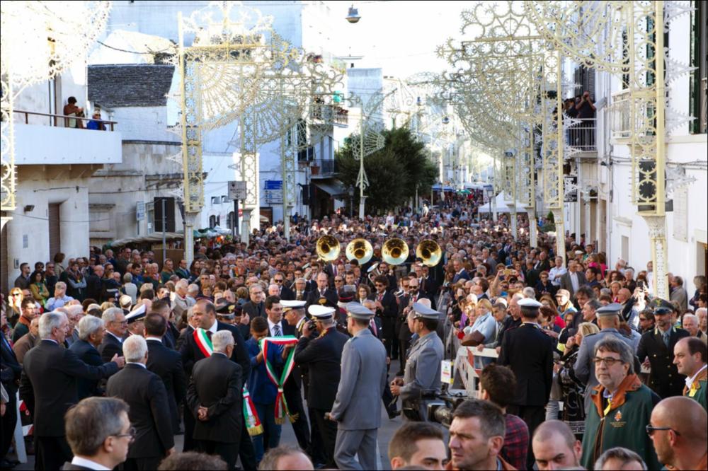 Lo scatto mostra devoti e pellegrini della città di Alberobello e non solo, accorsi per fare festa al passaggio dei musicisti della banda del paese che allietano la Festa in onore dei Santi Patroni Cosma e Damiano con concerti per le vie cittadine.
La Festa Patronale dei Santi Medici Cosma e Damiano, istituita il 12 aprile 1854, si celebra ad Alberobello, in provincia di Bari, dal 25 al 28 settembre.
Il 25 settembre si apre con la tradizionale fiera dell’antico bestiame e degli utensili agricoli. Il 26 settembre si celebra la festa liturgica. Il 27 settembre è dedicato ai pellegrini: la giornata inizia con la prima Messa alle ore 4.00, seguita dalla solenne processione mattutina alle ore 11.00. Il 28 settembre si concludono i festeggiamenti con ulteriori celebrazioni re...