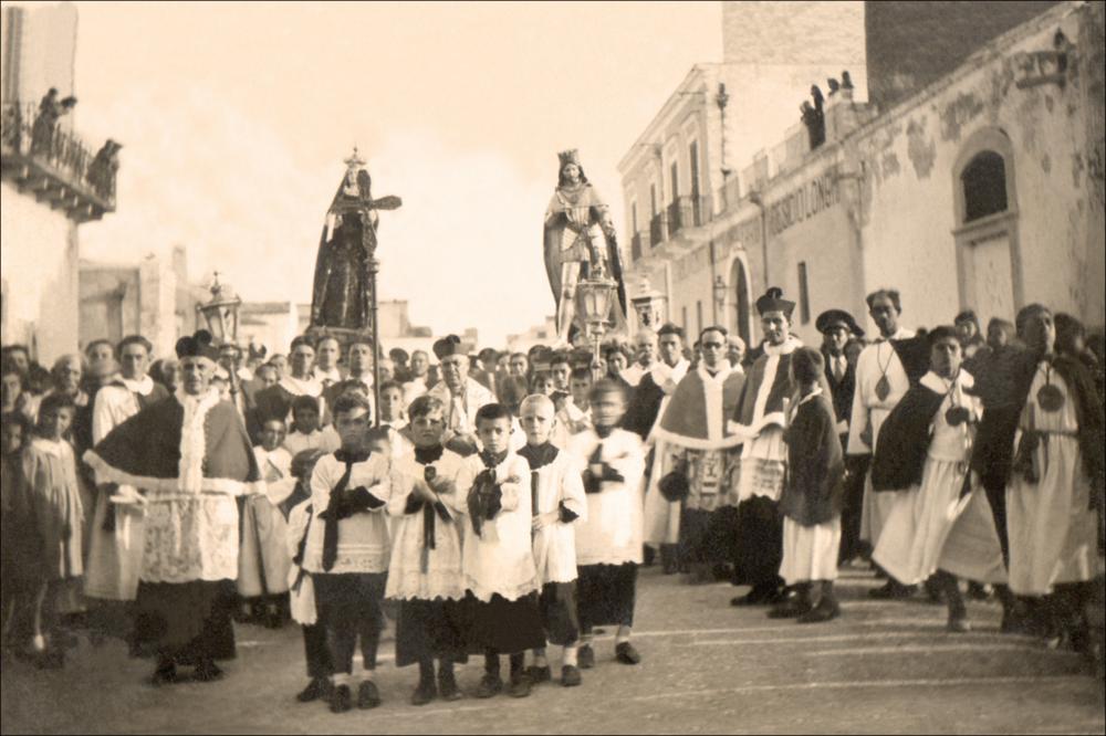 <div class="ql-editor"><div>La fotografia storica, di anno non definito, documenta un momento della processione patronale dedicata a San Ferdinando Re nelle strade di <strong>San Ferdinando di Puglia</strong>. Nell’immagine si riconosce il corteo religioso composto dal parroco e da altri sacerdoti, accompagnati da un gruppo numeroso di chierichetti che partecipano alla solenne celebrazione.</div><div>Al centro della scena compaiono i simulacri processionali della <strong>Madonna </strong>e di<strong> San Ferdinando Re</strong>, portati tra i fedeli durante il rito pubblico di devozione. Accanto al clero è visibile anche la presenza della confraternita: i partecipanti indossano abiti cerimoniali tipici delle confraternite cattoliche, con tuniche bianche, mante...