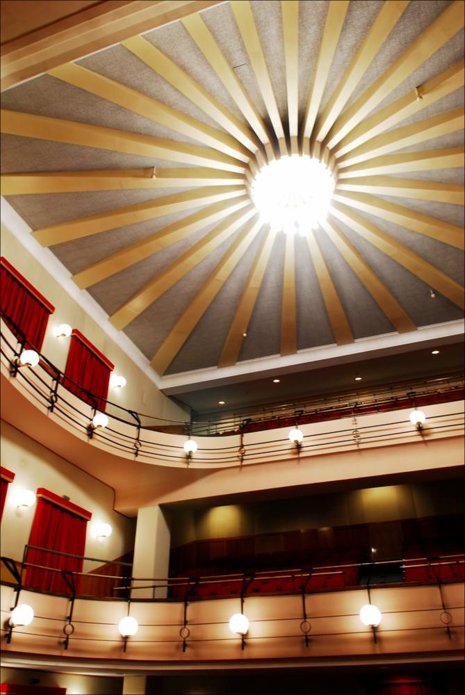 L'immagine è parte della campagna fotografica realizzata dalla fotografa Margherita Mirabella per Teatro Pubblico Pugliese, che ha ripreso nei suoi scatti le eleganti architetture e i rifiniti dettagli di alcuni dei più bei teatri pugliesi e alcuni degli spettacoli teatrali portati in scena in questi ultimi anni. L'autrice cattura il prospetto principale di Teatro Saverio Mercadante, situato in Piazza Giacomo Matteotti a Cerignola. Oggi gestito in collaborazione con il Teatro Pubblico Pugliese, la struttura teatrale fu inaugurata nel 1868. La storia del teatro inizia nel 1854, quando fu proposta la necessaria costruzione di un teatro cittadino. Dopo diverse proposte che risultarono piuttosto onerose, la scelta ricadde sull'architetto Leopoldo Vaccaro che presentò un pro...