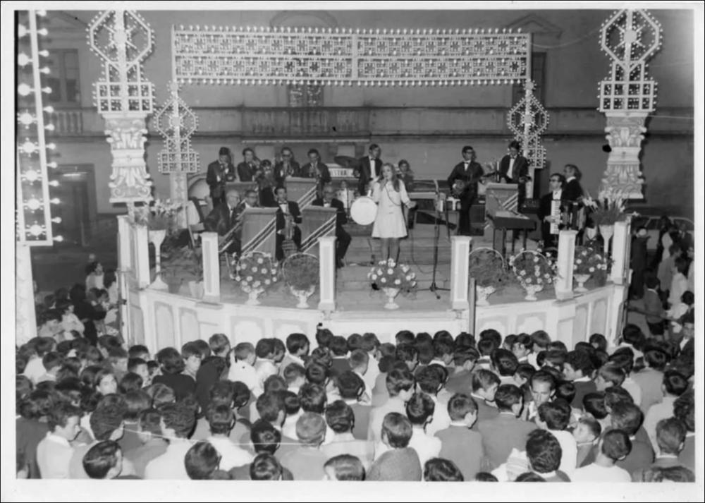 <div class="ql-editor"><div><strong>Foto storica del 1960. </strong>Scatto dall’alto che immortala l’esibizione in Piazza Municipio a Villa Castelli durante la Festa Patronale. La prospettiva permette di abbracciare con lo sguardo l’intero allestimento: il palco al centro della piazza, l’orchestra in piena esecuzione e il pubblico raccolto tutt’intorno. Il palco appare elegantemente decorato con composizioni floreali e impreziosito dalle luminarie che incorniciano la scena.</div><div><br></div><div>La Festa Patronale di <strong>Villa Castelli</strong> si svolge ogni anno dall’1 al 3 ottobre ed è uno degli appuntamenti più attesi e partecipati, non solo dai cittadini ma anche da tante persone provenienti dai paesi vicini. Con...
