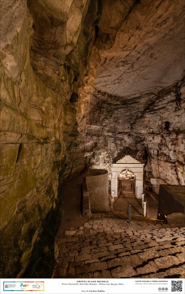 La grande caverna della Grotta di San Michele a Minervino Murge nel Parco Nazionale dell’Alta Murgia.
Il lento fluire dell’acqua di primitivi torrenti ha dato vita a questa meravigliosa cavità carsica risalente a due milioni di anni fa.  Ed è qui che circa mille anni fa è sorto questo affascinante luogo di culto dedicato a San Michele.
Un soffitto di pietra molto alto fa da cornice a questo ambiente straordinariamente suggestivo. Una scalinata monumentale in pietra conduce all’altare con la statua dell’Arcangelo.
L’immagine digitale fa parte della raccolta di fotografie prodotte da Carlos Solito per il Progetto digitale “Sillabario sotterraneo – Viaggio dantesco nelle Grotte di Puglia” – realizzato nell’ambito della LEGGE REGIONALE 30 dicembre 2020, n. 35 - art. 39 “Cav...