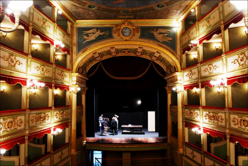L'immagine è parte della campagna fotografica realizzata dalla fotografa Margherita Mirabella per Teatro Pubblico Pugliese, che con i suoi scatti ha ripreso le eleganti architetture e i rifiniti dettagli di alcuni dei più bei teatri pugliesi e di alcuni spettacoli teatrali portati in scena in questi ultimi anni. La fotografia ritrae l'interno del Teatro Paisiello di Lecce, struttura teatrale progettata dall'architetto Oronzo Bernardini e sorta su una preesistente costruzione in legno risalente al XVIII secolo. Nel 1872 il teatro viene ricostruito in pietra e completato con delicati elementi decorativi affidati a maestranze napoletane. Insieme all'affresco della volta e agli elementi decorativi in stucco dorato, il piccolo teatro di Lecce conserva, nel foyer, due busti i...