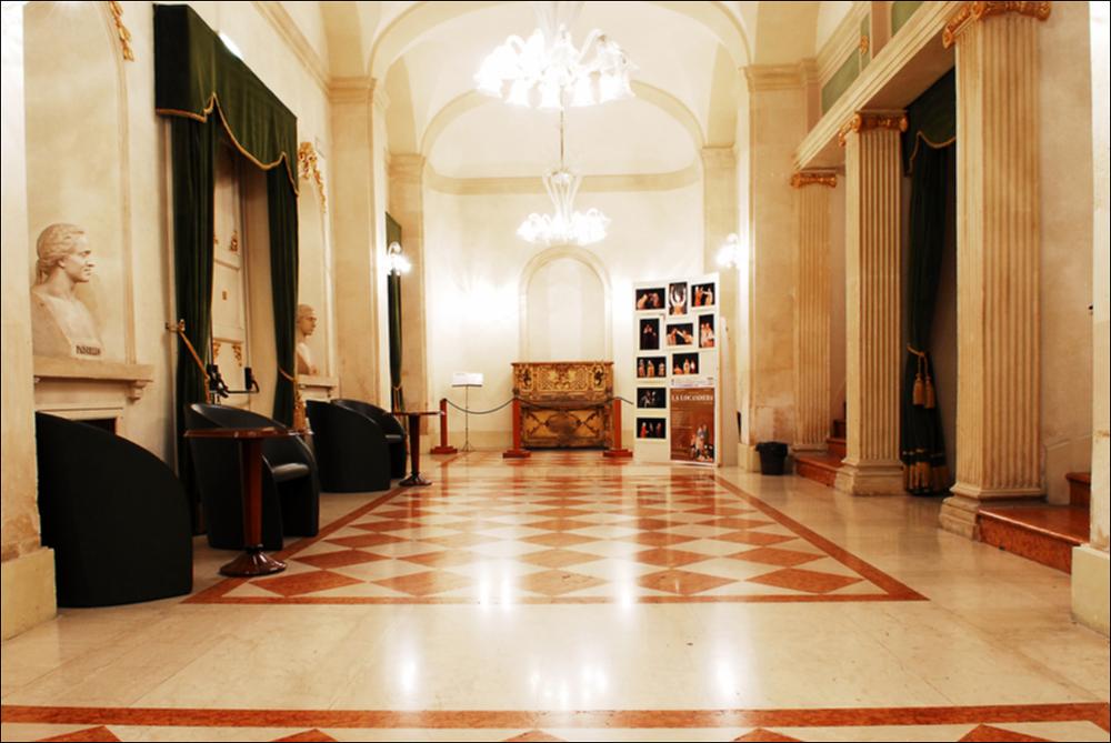 L'immagine è parte della campagna fotografica realizzata dalla fotografa Margherita Mirabella per Teatro Pubblico Pugliese, che con i suoi scatti ha ripreso le eleganti architetture e i rifiniti dettagli di alcuni dei più bei teatri pugliesi e di alcuni spettacoli teatrali portati in scena in questi ultimi anni. La fotografia ritrae l'area del foyer del Teatro Paisiello di Lecce, struttura teatrale progettata dall'architetto Oronzo Bernardini e sorta su una preesistente costruzione in legno risalente al XVIII secolo. Nel 1870 il teatro, dedicato al musicista tarantino Giovanni Paisiello, viene inaugurato con la rappresentazione dell'opera “Il ballo in maschera” di Giuseppe Verdi. Il grazioso teatro di Lecce conserva, nel foyer, due busti in marmo, uno dei quali raffigur...