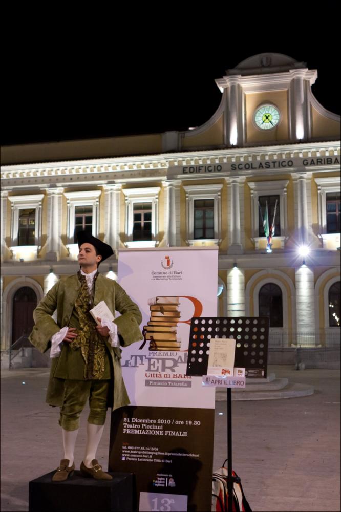 L'immagine digitale documenta una delle azioni di teatro urbano, messe in scena dall'Associazione Fibre Parallele, durante l'evento "Premio Letterario Città di Bari", dell'edizione 2010. La fotografia riprende l'attore che aspetta il suo pubblico davanti all'istituto scolastico “Garibaldi”, nel centro cittadino barese, per leggere le pagine del libro “Sono comuni le cose degli amici” di Matteo Nucci, edito da Ponte alle Grazie. Una storia tutta incentrata sull'amore di un figlio, Lorenzo, per il padre Leonardo, del quale dopo la sua morte, scopre la sua vera natura impulsiva e traditrice e cerca di capire se seguirne l'esempio vivendo di solitudine oppure dedicarsi faticosamente ad una vita fatta di autenticità ed affetti veri. L'edizione 2010 del Premio, alla sua XIII ...