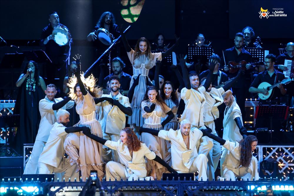 <div>Lo scatto ritrae il corpo di ballo della Notte della Taranta in una posa coreografica sul palco, con l’orchestra schierata alle loro spalle a occupare l’intera scena. L’immagine documenta uno dei momenti centrali dell’edizione 2022, evidenziando la sinergia tra danza e musica che caratterizza il festival salentino.</div>...