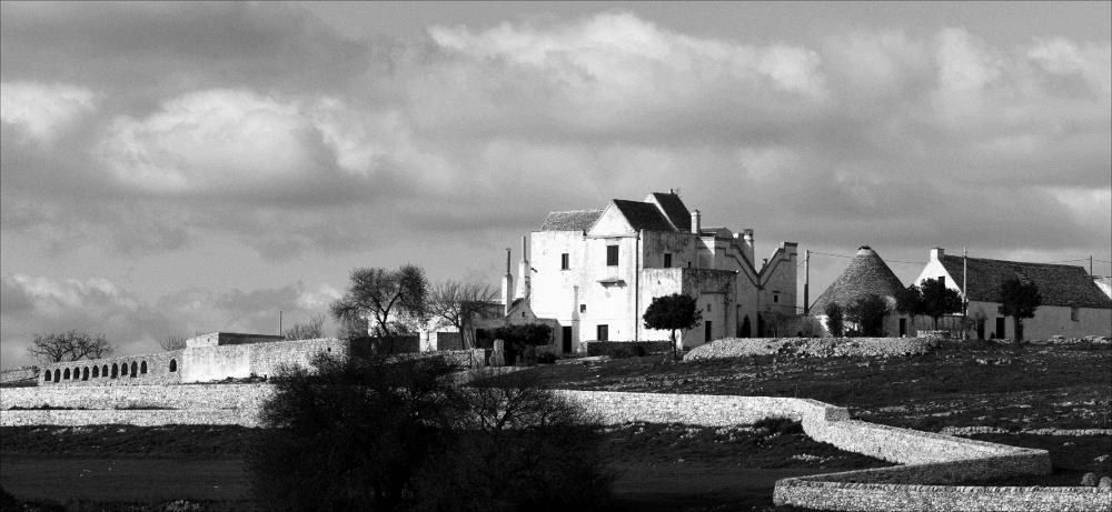 L'immaginein oggetto, scattata in bianco e nero e successivamente post prodotta, fa parte di una più ampia raccolta di fotografie realizzate per il progetto di censimento di 248 masserie pugliesi avviato da Giancarlo Chielli, professore del corso in Beni Culturali e Ambientali presso l'Accademia di Belle Arti di Bari. Il lavoro svolto ha dato vita al volume “Le masserie di Puglia come Bene Culturale” in cui sono presenti alcuni scatti di masserie pugliesi tra cui questa presa in esame. Lo scatto ci restituisce una veduta panoramica della masseria Sorresso, complesso architettonico risalente alla fine del XVIII secolo, nato in origine come abitazione rurale appartenente a Giannotto Sorresso e successivamente arricchito di ambienti e spazi di diversa funzione voluti dai p...