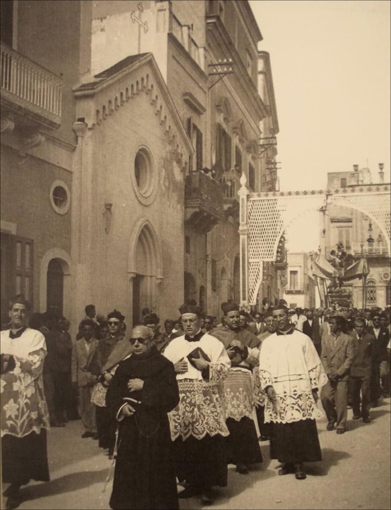 Scattata nel 1953, questa immagine storica ritrae un intenso momento della Festa Patronale dedicata a San Michele Arcangelo: la processione anima le strade di Massafra e, sullo sfondo, domina la scena la maestosa statua del Santo Patrono.
Il culto di San Michele, ufficializzato nel 1721 ma presente già da secoli, affonda le sue origini in antiche devozioni legate anche alla storica grotta carsica dedicata all’Arcangelo in zona Varcaturo, luogo di culto e testimonianza di riti e credenze tra epoche preclassiche e cristiane. Raffigurato come guerriero celeste, simbolo di protezione e giustizia, il santo è al centro di riti, opere d’arte, leggende e pratiche popolari che si tramandano di generazione in generazione, rafforzando il senso di appartenenza e coesione sociale.
N...