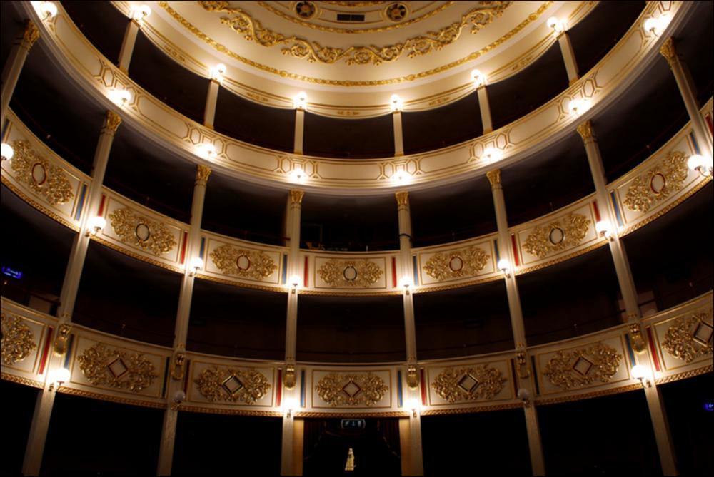 L'immagine è parte della campagna fotografica realizzata dalla fotografa Margherita Mirabella per Teatro Pubblico Pugliese, che con i suoi scatti ha ripreso le eleganti architetture e i rifiniti dettagli di alcuni dei più bei teatri pugliesi e di alcuni spettacoli teatrali portati in scena in questi ultimi anni. Lo scatto restituisce uno scorcio dei palchi del Teatro Comunale Tommaso Traetta di Bitonto. La struttura teatrale, intitolata solo nel 2005 al musicista bitontino Tommaso Traetta, fu voluta dalle famiglie notabili della città che, negli anni Trenta dell'800, reclamavano un attivo centro culturale dove poter sollazzare. I lavori di costruzione iniziarono nel 1835 e terminarono dopo soli tre anni, nel 1838. Nello stesso anno il teatro si aprì al pubblico con l'op...
