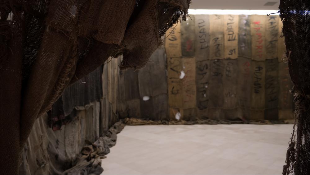 L’immagine presenta VOLI-NI, 1966-2021, una delle opere di Ibrahim Mahami, vincitore del Premio Pascali 2021 (XXIII edizione), in mostra presso la Fondazione Museo Pino Pascali di Polignano a mare dall’11 dicembre 2021 al 13 marzo 2022.
L'installazione è composta dai famosi sacchi di iuta assemblati dall’artista e dai suoi collaboratori e trasformati in drappeggi.
Mahama ricopre enormi monumenti con sacchi di juta, usati per il trasporto di carbone, sezioni di teloni impregnati d’olio e sigilli metallici, cuciti l’uno all’altro come fossero grandi arazzi “poveri”.
Il sacco di juta è il simbolo di un’economia fragile basata sulla produzione di cacao. Dietro ogni sacco di juta ci sono le vite delle persone che lo hanno maneggiato, timbrato, lacerato, rattoppato, trasporta...