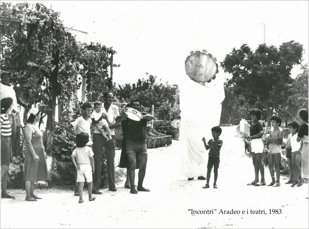 La fotografia in bianco e nero immortala un piccolo momento della rassegna “Aradeo e i Teatri” inaugurata nel suo primo anno con il titolo “Incontri”, un appuntamento con il teatro sperimentale che la compagnia Koreja, nata nel 1983 nel piccolo borgo salentino di Aradeo, portò avanti sin dall'anno della sua fondazione. La manifestazione fu organizzata dal circolo culturale “S.M. Ejzenstein” per colmare, attraverso la messa in scena di spettacoli di teatro e danza, la mancanza di confronto fra culture differenti molto sentita dalla cittadinanza di Aradeo. Nello specifico si osserva un gruppo di persone, molti bambini, contemplare meravigliati il “tamburello gigante”, strumento principe della pizzica tipico della tradizione salentina. La fotografia mette in luce come da o...