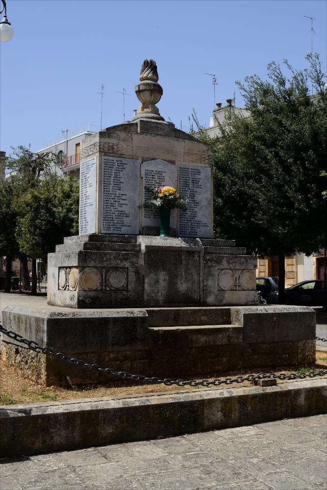 L'immagine digitale mostra una veduta d'insieme del Monumento ai Caduti di Castellana Grotte che nella parte centrale sui basamenti in pietra su tutti i lati riporta incisi i nomi dei caduti in guerra. L'opera, realizzata nel 1921 da Francesco Pinto (sul blocco scultoreo è riportata la firma), scultore originario di Castellana con studio a Firenze, fu poi rifatta nel 1948 quando Giovanni Laricchia sostituisce, con una iconografia completamente nuova, la scultura della patria in bronzo con una lampada votiva, che riprende stilisticamente quella in bassorilievo del retro.