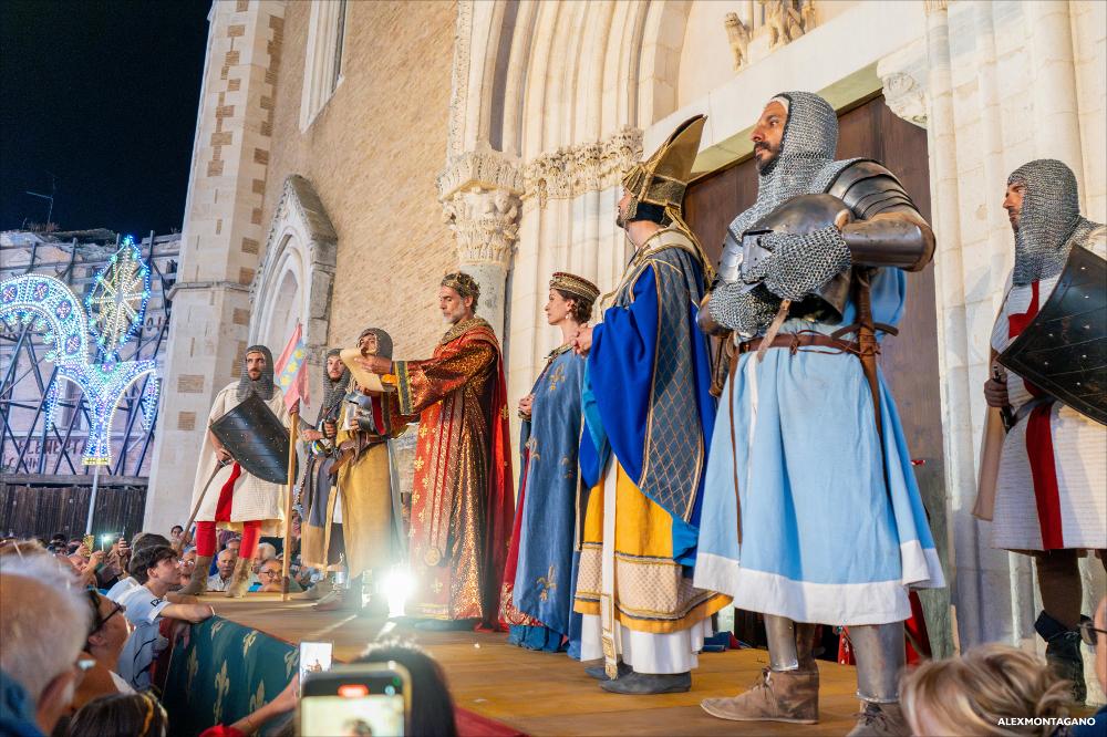 <div class="ql-editor"><div>La fotografia documenta la cerimonia di affidamento della Città a Santa Maria, atto conclusivo del Corteo Storico di Lucera. In Piazza Duomo, Sergio Muniz interpreta <strong>Carlo II d’Angiò</strong>, rievocando simbolicamente il gesto di consegna della città alla sua Patrona. La scena restituisce il valore rituale e identitario del momento, fulcro della Settimana Medioevale.</div><div><br></div><div>Il 13 agosto 2025, il <strong>Corteo Storico di Lucera</strong> ha animato le vie del centro, con partenza dalla Fortezza Svevo-Angioina e arrivo in piazza Duomo. La sfilata ha visto la partecipazione straordinaria di Sergio Muniz nelle vesti di re Carlo II d’Angiò e ha coinvolto oltre cinquecento figuranti –...