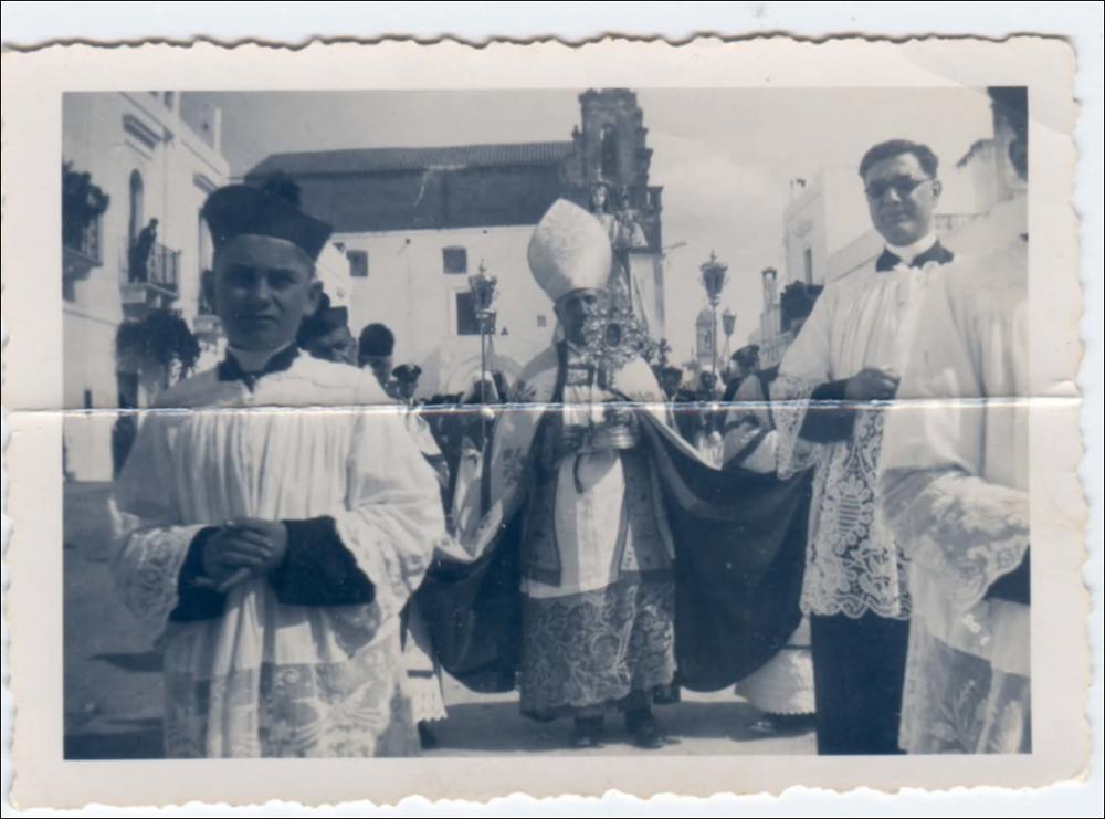 Nella fotografia della processione della Beata Vergine del Rosario del 1940 si coglie un momento di grande solennità. Al centro della scena, la statua della Madonna del Rosario avanza lentamente, portata dai fedeli su una base decorata, circondata da ceri, fiori e lacci d’onore.
Davanti al simulacro, a guidare la processione, si distingue il Vescovo: indossa un cappello bianco a punta (mitra), simbolo del suo ruolo ecclesiastico, e regge un reliquario, segno di autorità e protezione spirituale. La sua presenza sottolinea l’importanza liturgica della cerimonia, conferendo solennità e continuità al culto della Patrona.<div><br>La Festa Patronale della Beata Vergine del Santissimo Rosario di Montemesola (TA) è uno degli appuntamenti più sentiti e identitari della comunità....