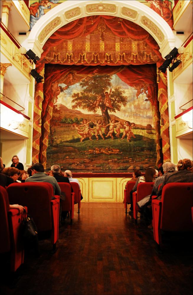L'immagine è parte della campagna fotografica realizzata dalla fotografa Margherita Mirabella per Teatro Pubblico Pugliese, che ha ripreso nei suoi scatti le eleganti architetture e i rifiniti dettagli di alcuni dei più bei teatri pugliesi e alcuni degli spettacoli teatrali portati in scena in questi ultimi anni. Siamo nella platea del piccolo ed elegante Teatro Comunale di Mola di Bari, intitolato al suo più celebre cittadino, il compositore Niccolò Van Westerhout. Iniziati nel 1888 per volere dell'Amministrazione comunale, i lavori per la sua edificazione si conclusero nel 1896; il teatro era pronto così ad ospitare sul suo palco il 18 aprile l'opera “Doña Flor”, che Van Westerhout dedicò a Mola, la sua città nativa. Scene e quinte furono dipinte dal barese Domenico B...