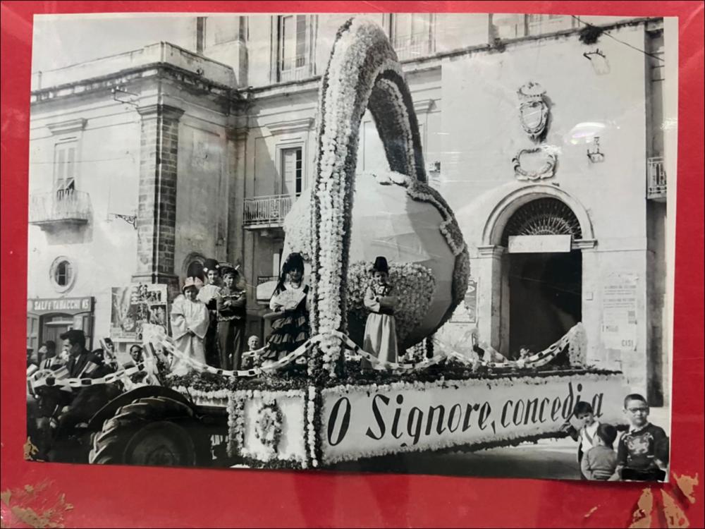 Questa foto storica degli anni ’60 mostra uno dei tradizionali carri realizzati per la "Sfilata dei carri votivi". La sfilata dei carri devozionali (o votivi) addobbati, che rievocano la leggenda del ritrovamento del crocifisso da parte dei contadini, è la principale celebrazione patronale della Festa del SS. Crocifisso di Auricarro a Palo del Colle e viene organizzata ancora oggi nel mese di maggio. I carri di Santa Croce sono strutture artistiche addobbate con forme di carta pesta, fiori e luminarie, rappresentanti temi religiosi, trainati da buoi, cavalli o trattori e realizzati a cura di associazioni, privati o scuole.
La Festa Patronale del SS. Crocifisso di Auricarro, istituita nel 1853, si celebra in due momenti diversi, il 3 Maggio e la terza settimana di settem...