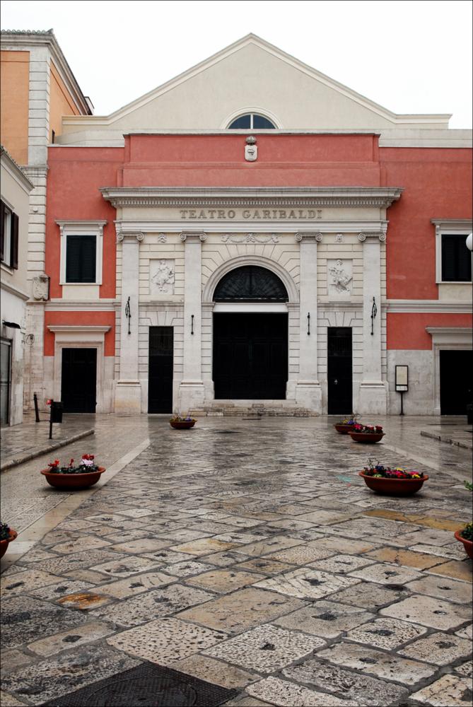 L'immagine è parte della campagna fotografica realizzata dalla fotografa Margherita Mirabella per Teatro Pubblico Pugliese, che ha ripreso nei suoi scatti le eleganti architetture e i rifiniti dettagli di alcuni dei più bei teatri pugliesi e alcuni degli spettacoli teatrali portati in scena in questi ultimi anni. Con una bella prospettiva centrale, l'autore cattura il prospetto principale di Teatro Garibaldi, situato nel borgo antico di Bisceglie tra Piazza Vittorio Emanuele II e Piazza Regina Margherita di Savoia. La sua costruzione fu voluta dal governo regio sabaudo, quando fu deliberata la decisione di trasformare gli antichi cameroni della “Polveriera”, dove sorgeva l'antico teatro cittadino costruito sugli ambienti ricavati da uno dei bastioni della cinta muraria....