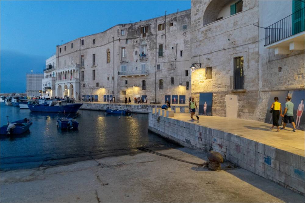 Lo scatto mostra l’immersione della mostra nelle attività quotidiane della città di Monopoli, partecipativa durante il festival PhEST. Le mura sono allestite con gli scatti di Mustafa Sabbagh (Amman, Giordania, 1961) del progetto “Corpus fugit”. I soggetti sono giovani ragazzi e ragazze che, immortalati su uno sfondo scuro, mettono in luce la loro individualità e la dimensione reale delle stampe aumenta il coinvolgimento dell’osservatore che si muove in questa “folla” di ragazzi....