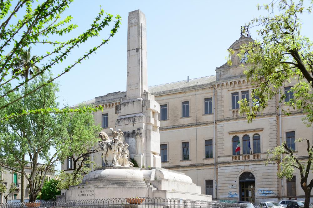 L'immagine digitale mostra il Monumento ai Caduti a Martina Franca. Opera dello scultore pugliese Antonio Bassi il monumento, sito in Piazza Vittorio Veneto, presenta un maestoso stilobate su cui poggia il grande obelisco. Nello specifico la fotografia riprende un dettaglio della parte frontale del monumento in marmo bianco di Carrara, dove vi è collocato un gruppo scultoreo in pietra di Trani, definito dallo stesso autore degli “Eroi”, raffigurante una scena di battaglia che vede protagonisti un gruppo di soldati e due cavalli nell'atto di impennarsi. Nella parte anteriore dell'obelisco fu apposta la semplice scritta “Ai suoi eroi, Martina Franca”. Nella parte posteriore del pilastro è collocato un altorilievo in pietra definito “dell'Olocausto” raffigurante un giovane...