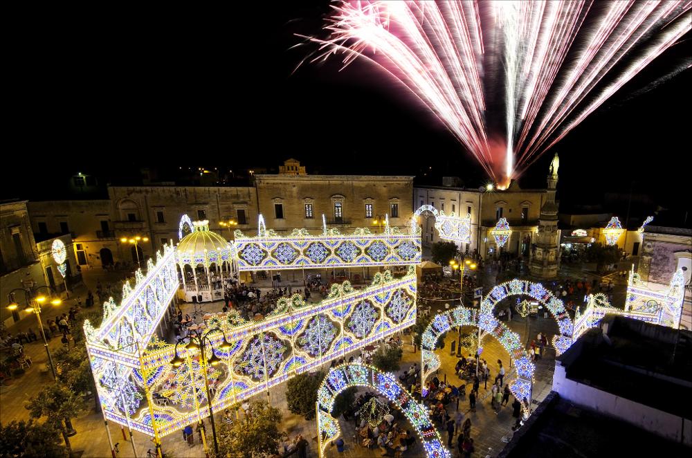 La piazza principale di Vernole, comunemente identificata con Piazza Vittorio Veneto rappresenta il cuore storico, religioso e sociale del centro salentino. Luogo di incontro quotidiano e di aggregazione comunitaria, essa assume un ruolo centrale durante le principali ricorrenze civili e religiose.
In particolare, ospita i solenni festeggiamenti in onore dei Santi Gioacchino e Anna, patroni del paese, celebrati il 25 e 26 luglio. In tali giornate la piazza si trasforma scenograficamente: le tradizionali luminarie artistiche incorniciano lo spazio urbano, la cassa armonica accoglie le esibizioni bandistiche e la partecipazione popolare anima ogni angolo del centro storico.
Momento culminante delle celebrazioni sono gli spettacoli pirotecnici serali, che illuminano il cie...