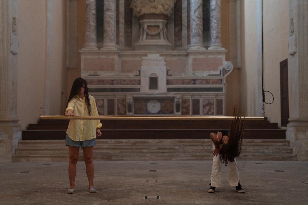 <div>La foto ritrae un momento dell’installazione performativa <strong>“Senza Titolo”</strong> di <strong>Romeo Castellucci</strong>, presentata il <strong>16 luglio 2024</strong> nella <strong>Chiesa di San Francesco della Scarpa</strong> per il <em>Teatro dei Luoghi Festival Internazionale</em>. L’opera propone gesti ripetuti, suoni e movimenti essenziali, creando un’esperienza immersiva e riflessiva nello spazio sacro. La scena invita il pubblico a osservare attentamente la materialità dei corpi e dei suoni, trasformando la chiesa in un luogo di percezione intensa e contemplativa.</div>...