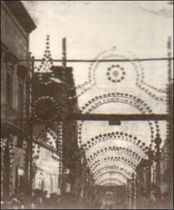 Una fotografia storica della festa patronale di fine Ottocento – inizi Novecento. L’immagine mostra le vie principali del paese ornate da luminarie ad acetilene, costituite da strutture lignee decorate e impreziosite da piccole fiammelle vive, alimentate da bruciatori che diffondevano una luce calda e tremolante. La fotografia seppiata mette in risalto il contrasto tra il buio della sera e i punti luminosi delle fiammelle, simbolo di progresso tecnico per l’epoca, ma anche della continuità del rito. Le luminarie ad acetilene rappresentavano infatti una novità tecnologica che, pur con mezzi semplici, trasformava lo spazio urbano in uno scenario festivo straordinario.<div><br>La Festa Patronale della Beata Vergine del Santissimo Rosario di Montemesola (TA) è uno degli app...