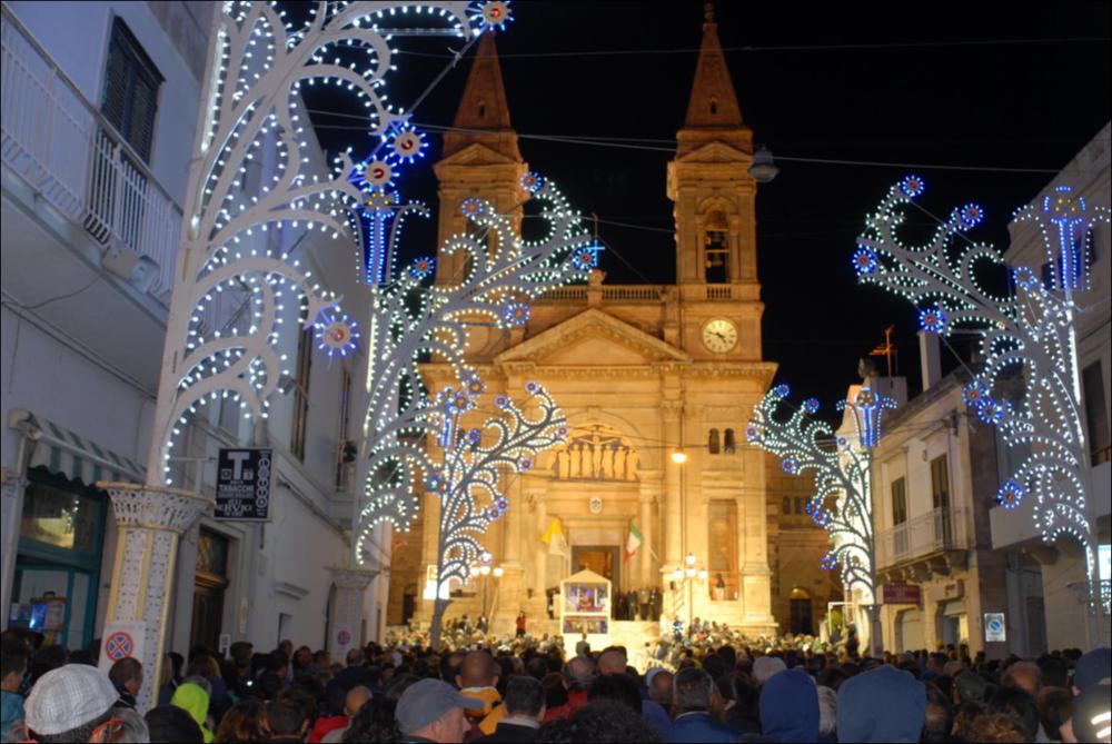 Lo scatto fotografico mostra la facciata della Chiesa Madre dei Santi Medici Cosma e Damiano che domina la via principale del paese gremita di fedeli e pellegrini che arrivano da ogni dove, accolti da una città illuminata a giorno da luci e colori sfavillanti.
La Festa Patronale dei Santi Medici Cosma e Damiano, istituita il 12 aprile 1854, si celebra ad Alberobello, in provincia di Bari, dal 25 al 28 settembre.
Il 25 settembre si apre con la tradizionale fiera dell’antico bestiame e degli utensili agricoli. Il 26 settembre si celebra la festa liturgica. Il 27 settembre è dedicato ai pellegrini: la giornata inizia con la prima Messa alle ore 4.00, seguita dalla solenne processione mattutina alle ore 11.00. Il 28 settembre si concludono i festeggiamenti con ulteriori cel...