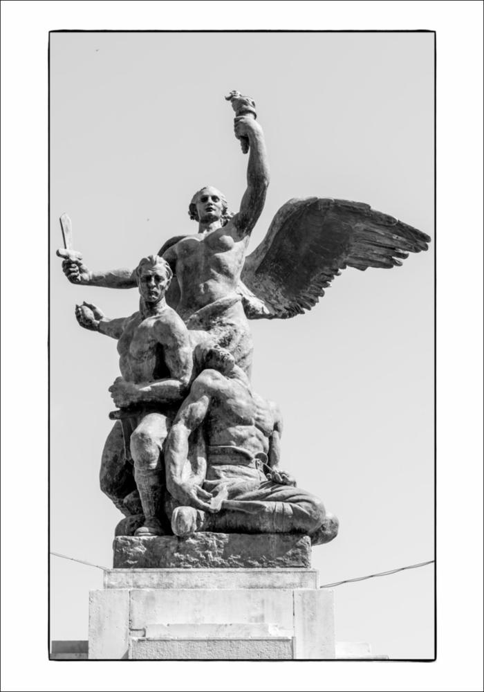 L'immagine in bianco e nero immortala il gruppo scultoreo del Monumento ai Caduti di Cassano delle Murge. Posizionato al centro di Piazza Dante Alighieri, è realizzato in bronzo, marmo e pietra come un'ara sul cui basamento sono riportate le lastre con incisioni dei concittadini caduti nelle due guerre mondiali. Degne di nota sulla sommità sono visibili le statue in bronzo, con al centro l'Allegoria della Vittoria, realizzate dalla Fonderia / Marmeria Chiurazzi su progetto artistico dello scultore Vitantonio De Bellis. L'artista è annoverato tra i casi più interessanti a livello pugliese, autore di altri monumenti pugliesi come quello di Rutigliano e Carbonara, tanto da poterlo definire uno specialista del genere, attività fortunata che porterà lo stesso artista a dedic...