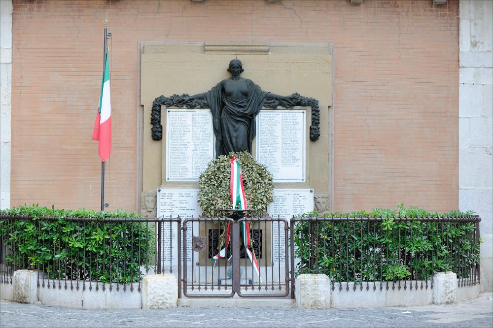 L'immagine digitale mostra un particolare del Monumento ai Caduti di Lucera, opera dello scultore toscano Giulio Passaglia che realizzò la lapide monumentale unendo materiali diversi come il bronzo, la pietra di Siena e la pietra di Trani. Collocata su una parete di Palazzo Venditti, nella centrale piazza Duomo, la lapide presenta una composizione con uno sfondo di pietra di Siena che mostra, lungo il perimetro, una serie di lavorazioni che creano una cornice ai nomi dei caduti in guerra intagliati nel ricordo ai posteri. Sotto la cornice è incisa l'epigrafe “Dulce et decorum pro patria mori”, verso ripreso dal libro terzo delle Odi di Orazio, da tradurre letteralmente “è dolce e dignitoso morire per la patria”. L'altorilievo bronzeo raffigura una donna alta e vigorosa,...