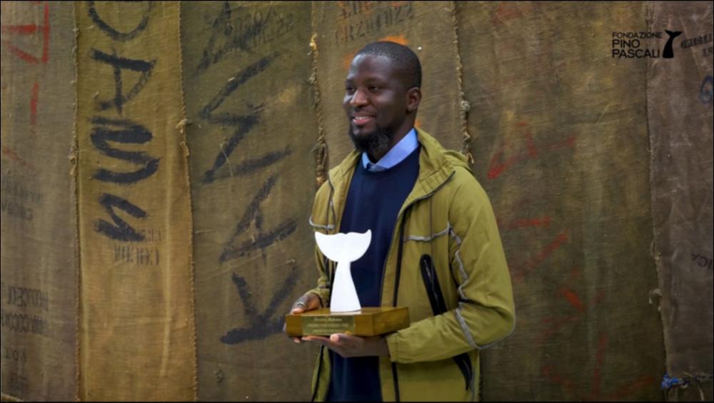 Questo breve video presenta l’opera di Ibrahim Mahama (Tamale, Ghana 1987), noto artista ghanese da diversi anni protagonista della scena internazionale, vincitore del Premio Pino Pascali 2021 – XXIII edizione, in mostra presso la Fondazione Museo Pino Pascali di Polignano a mare. Nel video sono presenti gli interventi dei membri della Commissione: il Presidente della Fondazione Museo Pino Pascali, Giuseppe Teofilo, la direttrice della Fondazione Museo Pino Pascali, Rosalba Branà, la direttrice della Fondazione Giuliani per l’arte contemporanea di Roma, Adrienne Drake, e lo storico dell’arte e curatore della Fondazione Pino Pascali, Nicola Zito. Segue una presentazione delle opere da parte dello stesso artista Ibrahim Mahama.
L’arte di Mahama è fatta di contaminazioni, ...
