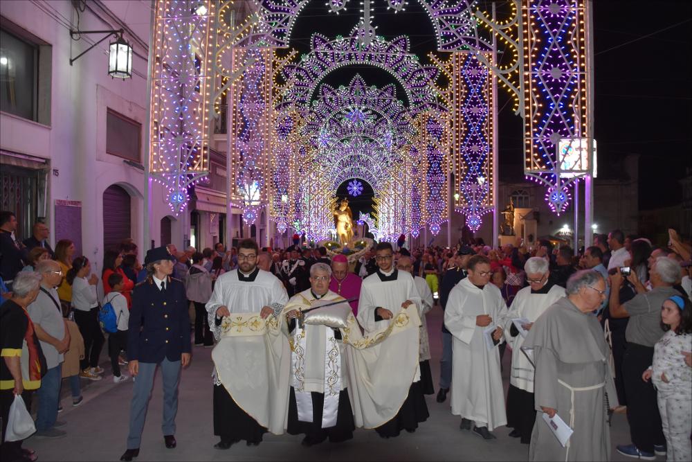 L’immagine cattura il suggestivo passaggio della Processione in onore di San Michele Arcangelo lungo le vie cittadine, illuminate da scintillanti luminarie che rischiarano il cielo della sera e accompagnate dalla gioiosa partecipazione di una folla di fedeli in festa.
In primo piano spicca la Sacra Spada, portata in processione per impartire la benedizione alla città e ai numerosi pellegrini raccolti dinanzi al Santo Patrono.
Il “Rito della Sacra Spada” si celebra il 29 settembre all’interno della Grotta del Santuario: durante la cerimonia il vescovo estrae la spada dalla statua, compiendo un gesto altamente simbolico che richiama la Parola di Dio, capace di discernere il bene dal male e di donare protezione e guarigione spirituale ai fedeli. La reliquia viene quindi po...