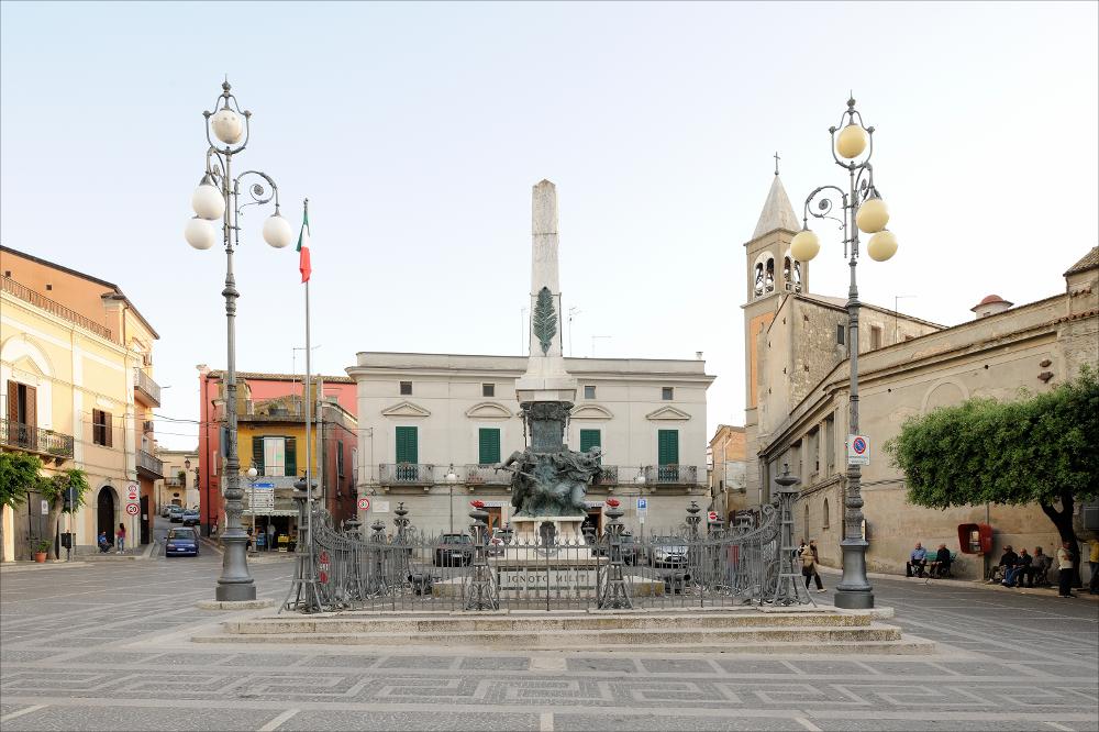 L'immagine digitale mostra il Monumento ai Caduti ad Ascoli Satriano, opera dello scultore bolognese Giulio Barberi. Racchiuso da una cancellata in ferro battuto, su un doppio gradone, vi è un blocco in marmo sul quale si erge un gruppo bronzeo, sul quale a sua volta si innalza un obelisco su cui sono state inserite delle foglie di palma in bronzo. Il gruppo scultoreo consta di due protagonisti: a sinistra un bersagliere è ritratto nel momento di avanzamento alla carica con moschetto e baionetta innestata, mentre sulla destra vi è un fante colpito che cade avendo tra le braccia la bandiera. I due personaggi sembrano sporgersi da una parete rocciosa. Sui lati dell'obelisco sono presenti delle targhe bronzee: una in facciata con lo stemma bronzeo del comune di Ascoli Satr...