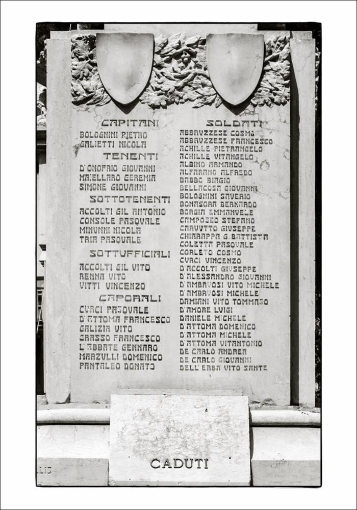 L'immagine è un dettaglio con le incisioni dei nomi dei caduti appartenente al Monumento ai Caduti di Conversano. Ubicato nella zona centrale della città al termine della Villa comunale, il monumento fu concepito come una stele in pietra onoraria denominata col termine "Piramide in travertino", sulla cui sommità svetta una stella e fu realizzato dall'artista pugliese originario di Rutigliano Vitantonio De Bellis, autore di diversi monumenti sul territorio che divenne uno specialista del genere, un'attività fortunata che lo porterà poi a dedicarsi, negli anni '60-'70, al restauro e al reintegro dei suoi stessi monumenti. La fotografia è stata esposta nel Salone degli Affreschi del Palazzo Ateneo di Bari per la mostra fotografica “La memoria della Grande Guerra in Puglia”...