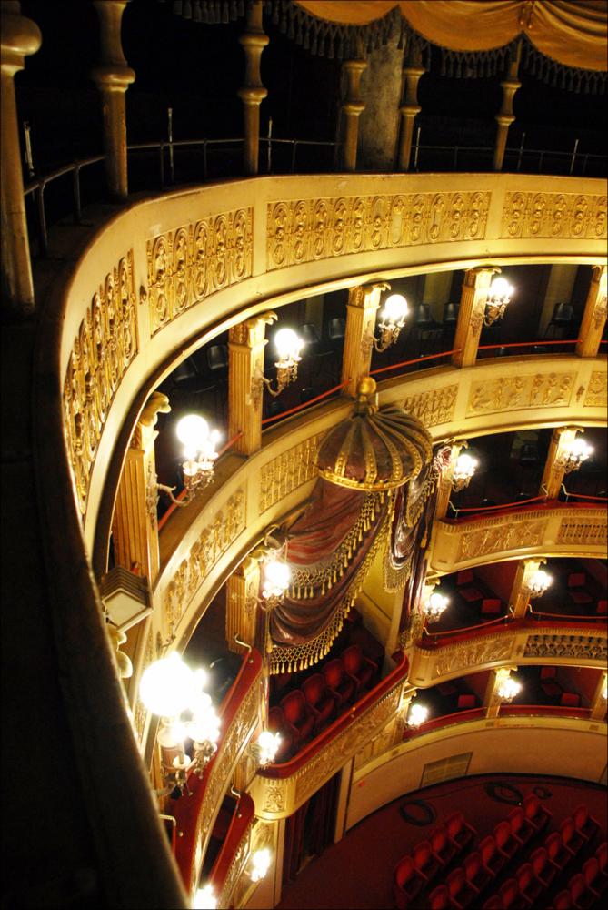 L'immagine è parte della campagna fotografica realizzata dalla fotografa Margherita Mirabella per Teatro Pubblico Pugliese, che con i suoi scatti ha ripreso le eleganti architetture e i rifiniti dettagli di alcuni dei più bei teatri pugliesi e di alcuni spettacoli teatrali portati in scena in questi ultimi anni. La foto restituisce uno scorcio degli eleganti palchi, finemente decorati, del Teatro Comunale Niccolò Piccini, la più antica struttura teatrale della città di Bari. I lavori di costruzione del teatro furono avviati nel 1836 su progetto dell'architetto Antonio Niccolini e ultimati nel 1854; nello stesso anno fu inaugurato con l'allestimento del Poliuto di Gaetano Donizetti e nel 1855 venne dedicato al compositore barese Niccolò Piccini. Il Piccinni è il quarto t...