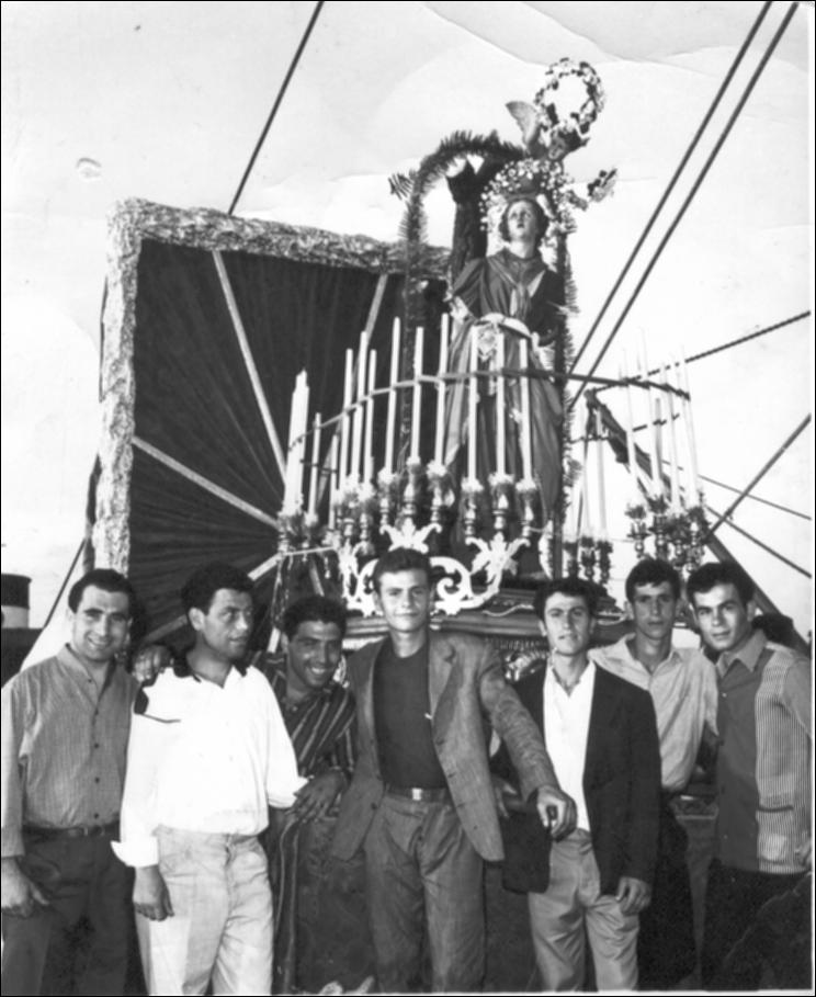 Fotografia storica degli anni 60 raffigurante un gruppo di uomini, presumibilmente appartenenti al comitato organizzatore, posizionati dinanzi alla statua di Santa Cristina. Il simulacro appare solennemente allestito con candele votive e panneggi decorativi, secondo l’uso tradizionale delle celebrazioni patronali.

"Santa Cristina sul pontile" è tra i momenti più suggestivi della festa patronale vi è la presenza della statua di Santa Cristina sul pontile del porto di Gallipoli. Il simulacro viene condotto in prossimità del mare, luogo identitario della città, dove incontra simbolicamente la comunità dei pescatori e dei lavoratori del mare.
La Santa, rivolta verso le acque, richiama il legame profondo tra Gallipoli e il suo porto, cuore economico e sociale della città.

...