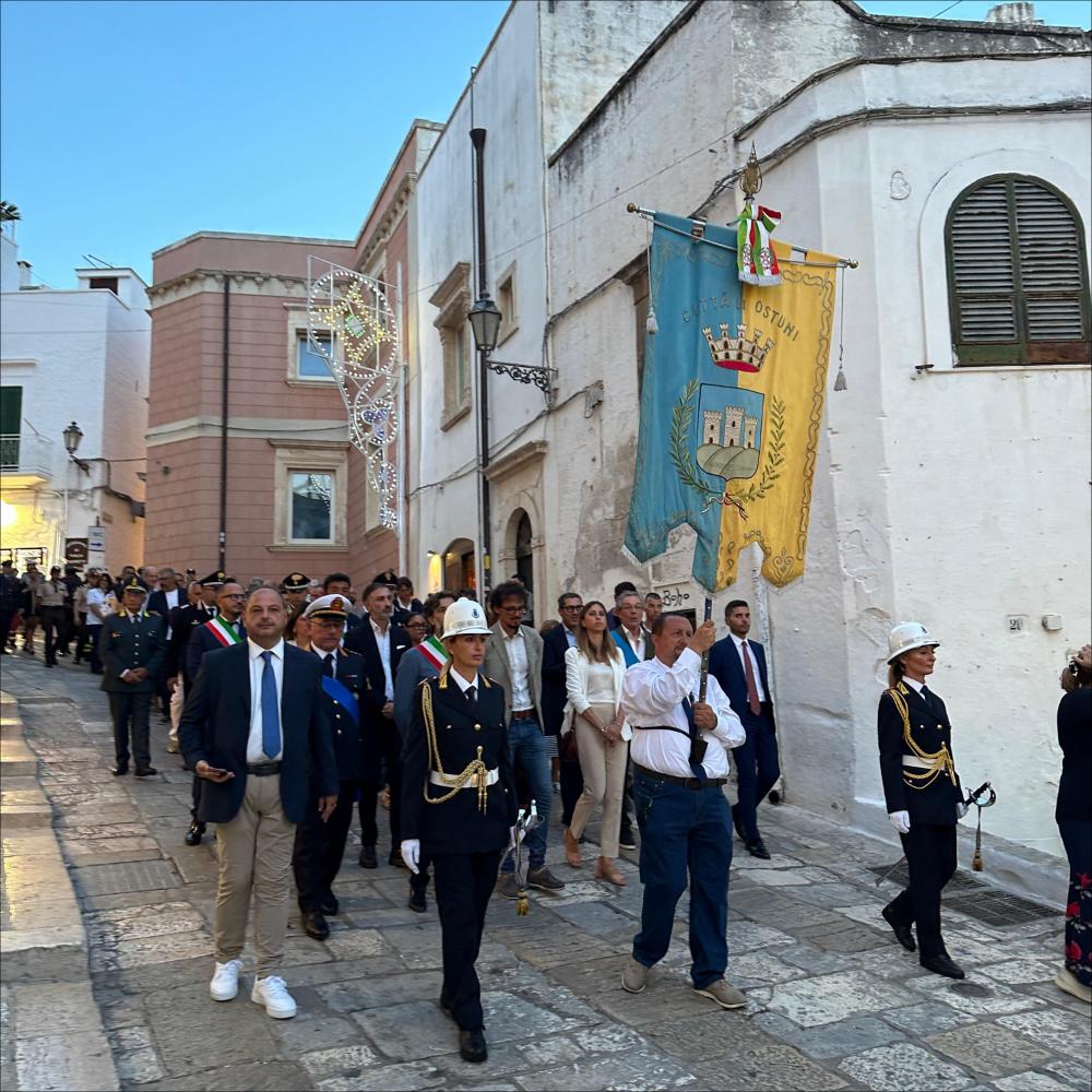 <div class="ql-editor"><div>Nell’immagine, in primo piano si distingue lo <strong>stendardo della città di Ostuni</strong>, simbolo civico che apre la processione per le vie della città in occasione dei festeggiamenti in onore di Sant'Oronzo. Subito dietro, si possono vedere i <strong>rappresentanti del Comune</strong>, che partecipano ufficialmente al corteo accanto ai cavalieri della Cavalcata e ai fedeli.</div><div><br></div><div>A Ostuni, ogni anno dal 25 al 27 agosto, si rinnova uno degli appuntamenti più sentiti dalla comunità: la Cavalcata in onore di Sant'Oronzo, solenne processione equestre che unisce fede, tradizione e identità cittadina. Attualmente la processione e la Cavalcata sono rese possibili grazie alla sinergia tra la Pa...